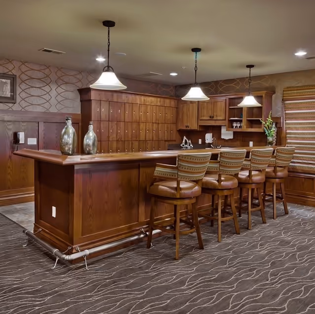 Wood-paneled lounge bar with four upholstered barstools, pendant lights, and cabinets in a carpeted room.