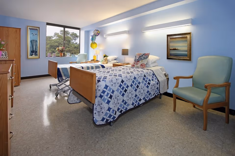 A bright and clean room with two hospital-style beds covered in blue and white patterned quilts. Each bed has pillows and a bedside table with lamps. There is a large window with a view of trees, a wooden dresser, a framed picture on the wall, and a comfortable armchair with wooden arms. Balloons and flowers decorate the room, creating a welcoming atmosphere.