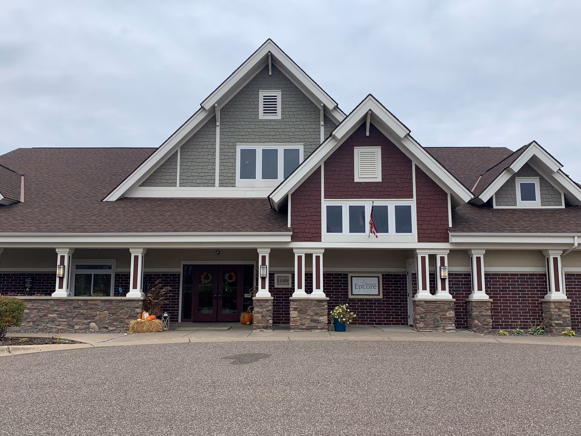 Front exterior view of Encore Assisted Living and Memory Care at Champlin building with a peaked roof, stone and brick facade, and a covered entrance with columns. There are some fall decorations near the entrance and an American flag hanging above the door.