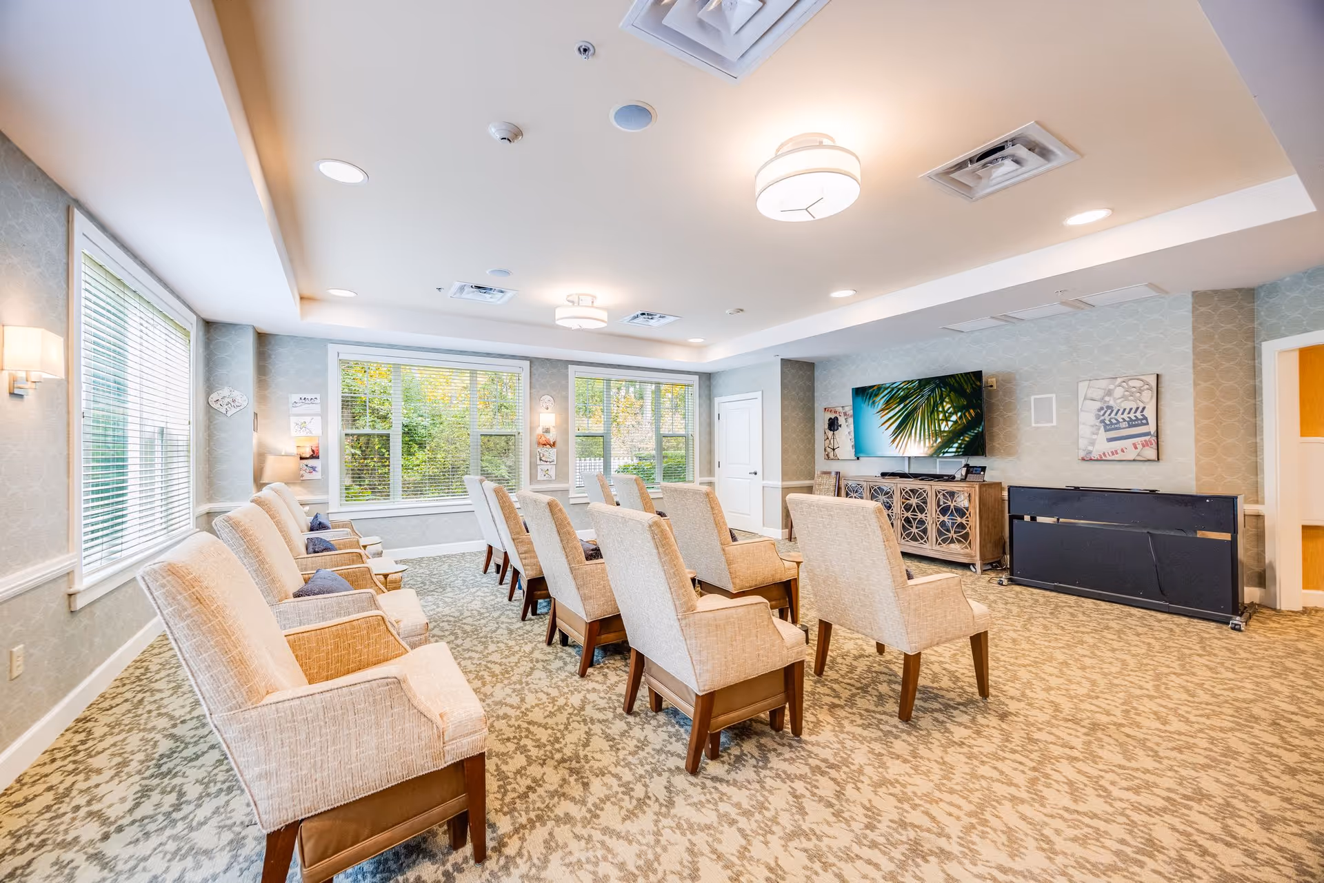 Bright common lounge with rows of upholstered chairs facing a wall-mounted TV and large windows.