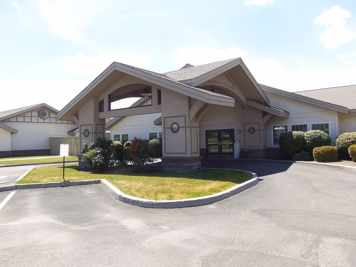 Front exterior entrance of a single-story senior care facility with a covered porte-cochère, landscaping, and a parking area.