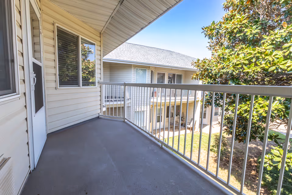 Covered second-floor balcony with a metal railing overlooking a courtyard, neighboring building, and trees.