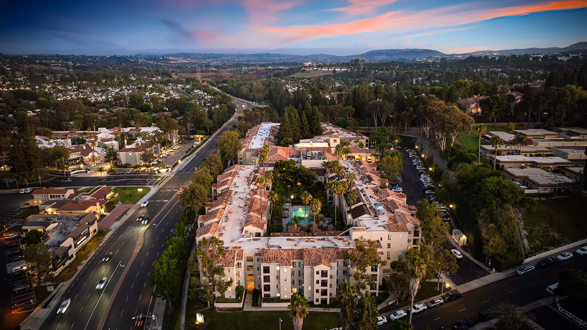 Aerial view of The Ivy at Wellington senior living facility at dusk, showing a large U-shaped building with a central courtyard featuring a swimming pool, surrounded by trees and parking areas, with a road and residential neighborhood nearby under a colorful sky.