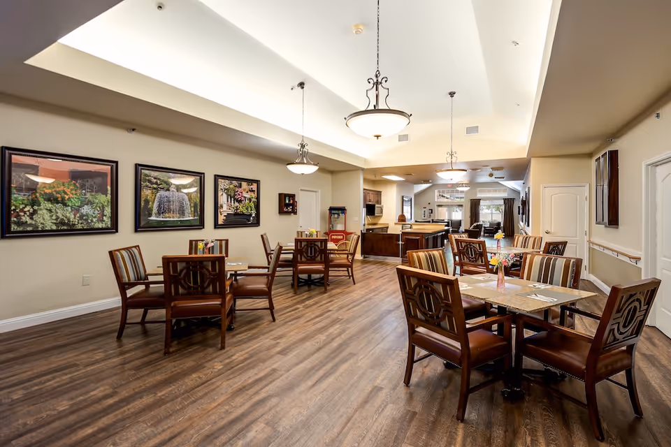 A spacious dining room with several wooden tables and chairs arranged neatly. The room features wood flooring, beige walls, and three hanging light fixtures. There are framed pictures of nature scenes on the wall to the left. In the background, there is a kitchen area with dark wood cabinets and a countertop, and further back, a living room area is visible.