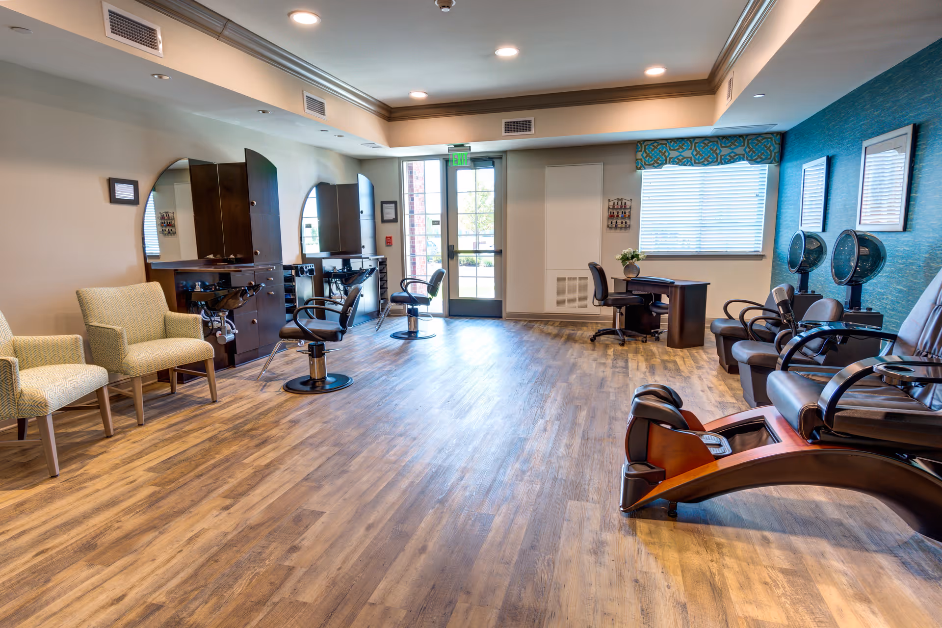 A spacious salon area in a senior living facility with wooden flooring, two styling chairs in front of large round mirrors, two beige armchairs, a desk with a chair near a window with blinds, and three brown leather pedicure chairs with foot baths and hair dryers mounted on the wall.