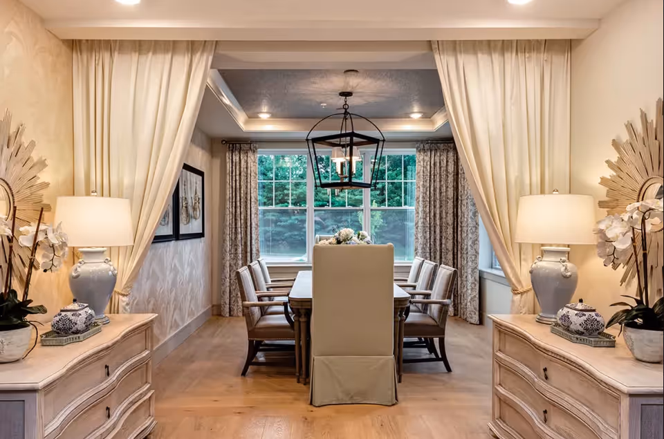 A well-lit dining room with a long wooden dining table surrounded by eight upholstered chairs. The room features large windows with patterned curtains, two side tables with matching lamps and decorative items, and light-colored walls with elegant wallpaper. A modern chandelier hangs above the table, and the floor is made of light wood.