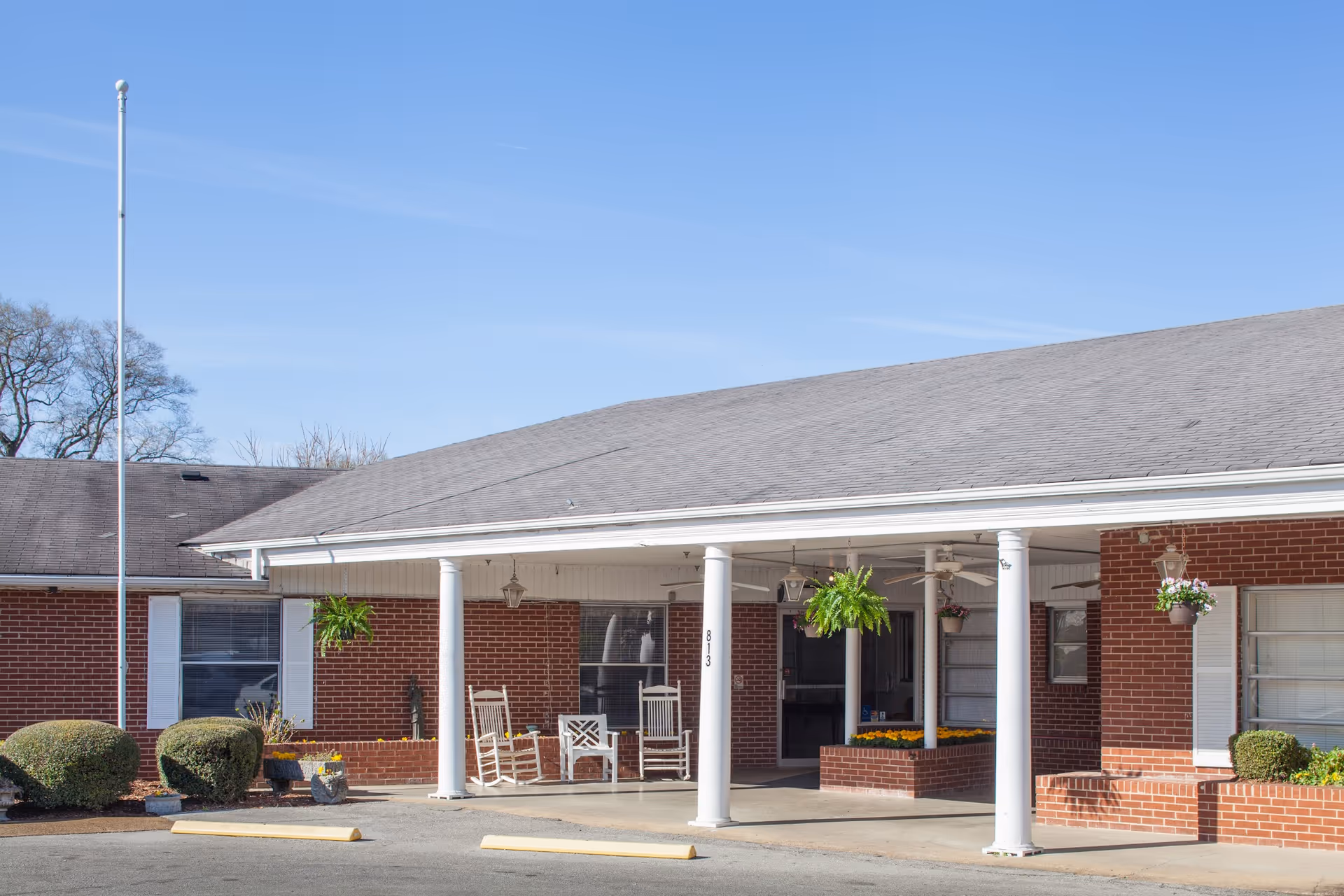 Front entrance of a brick senior living building with white columns, rocking chairs, and hanging plants.
