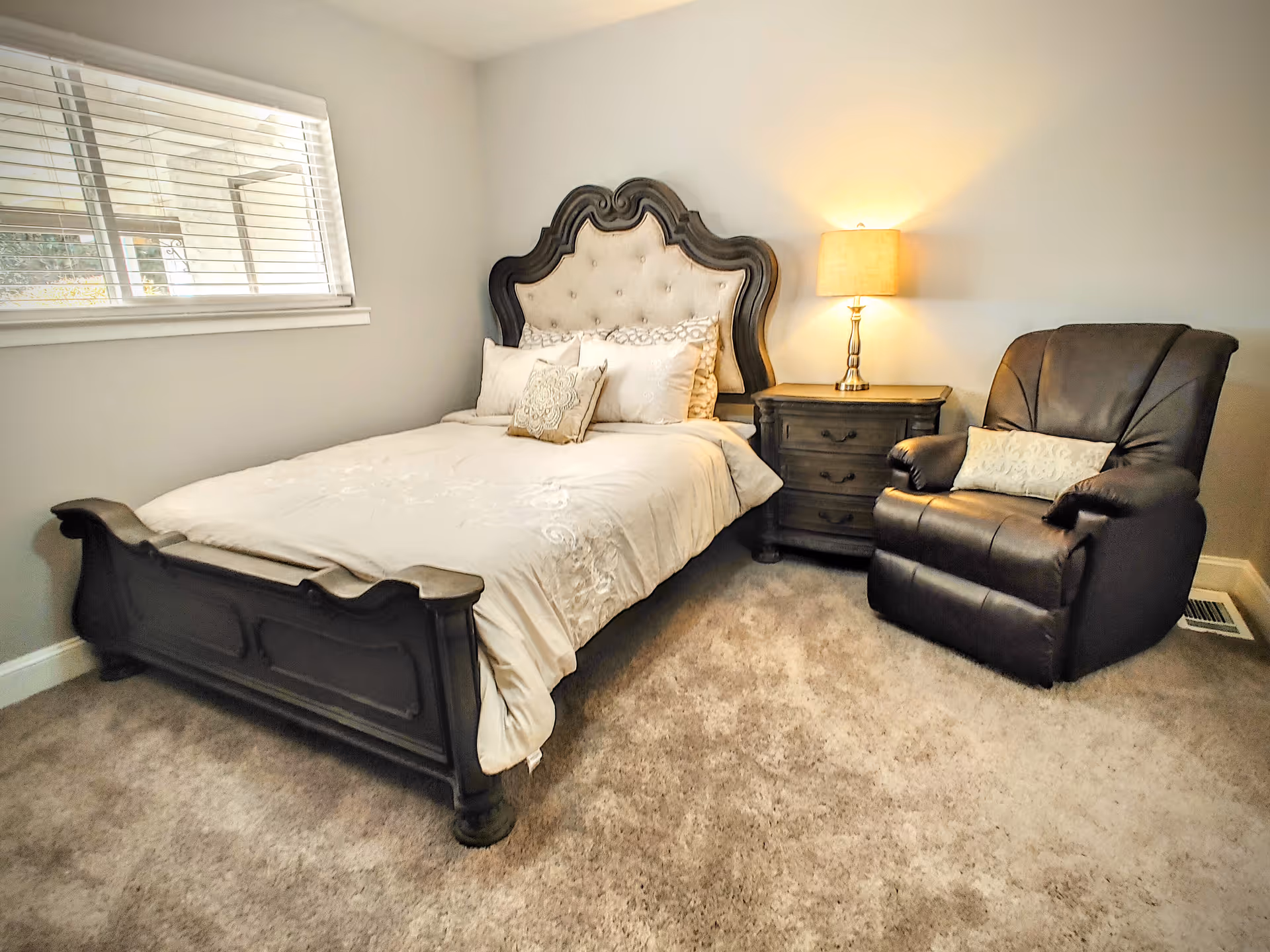 A cozy bedroom with a dark wooden bed frame featuring an upholstered headboard, a neatly made bed with white bedding and decorative pillows, a wooden nightstand with a lamp, and a dark brown leather recliner chair with a small pillow, all set on a carpeted floor under a window with white blinds.