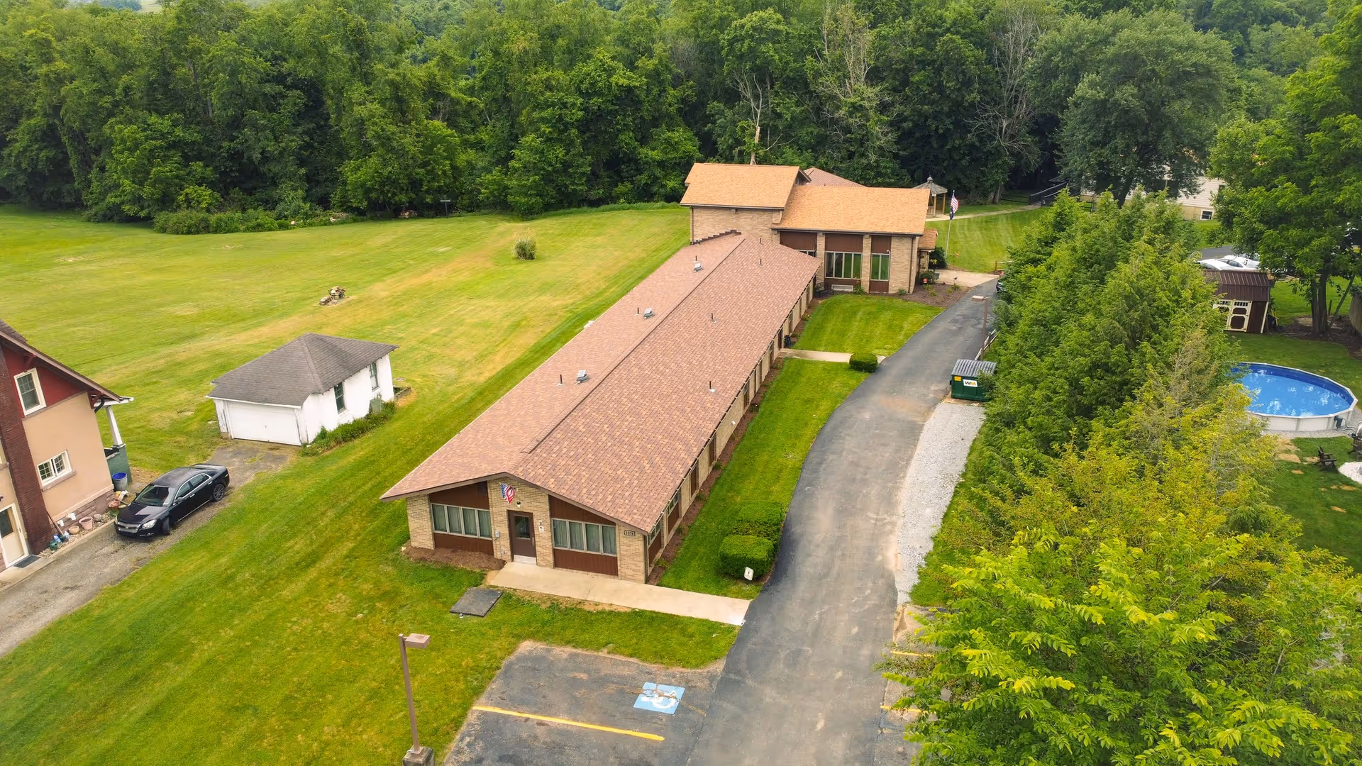 Aerial view of Vida Springs Senior Living and Personal Care Home showing a long, single-story building with a brown roof surrounded by green lawns and trees. There is a driveway alongside the building, a small parking area with a handicap parking spot, and a round above-ground pool visible in the background.