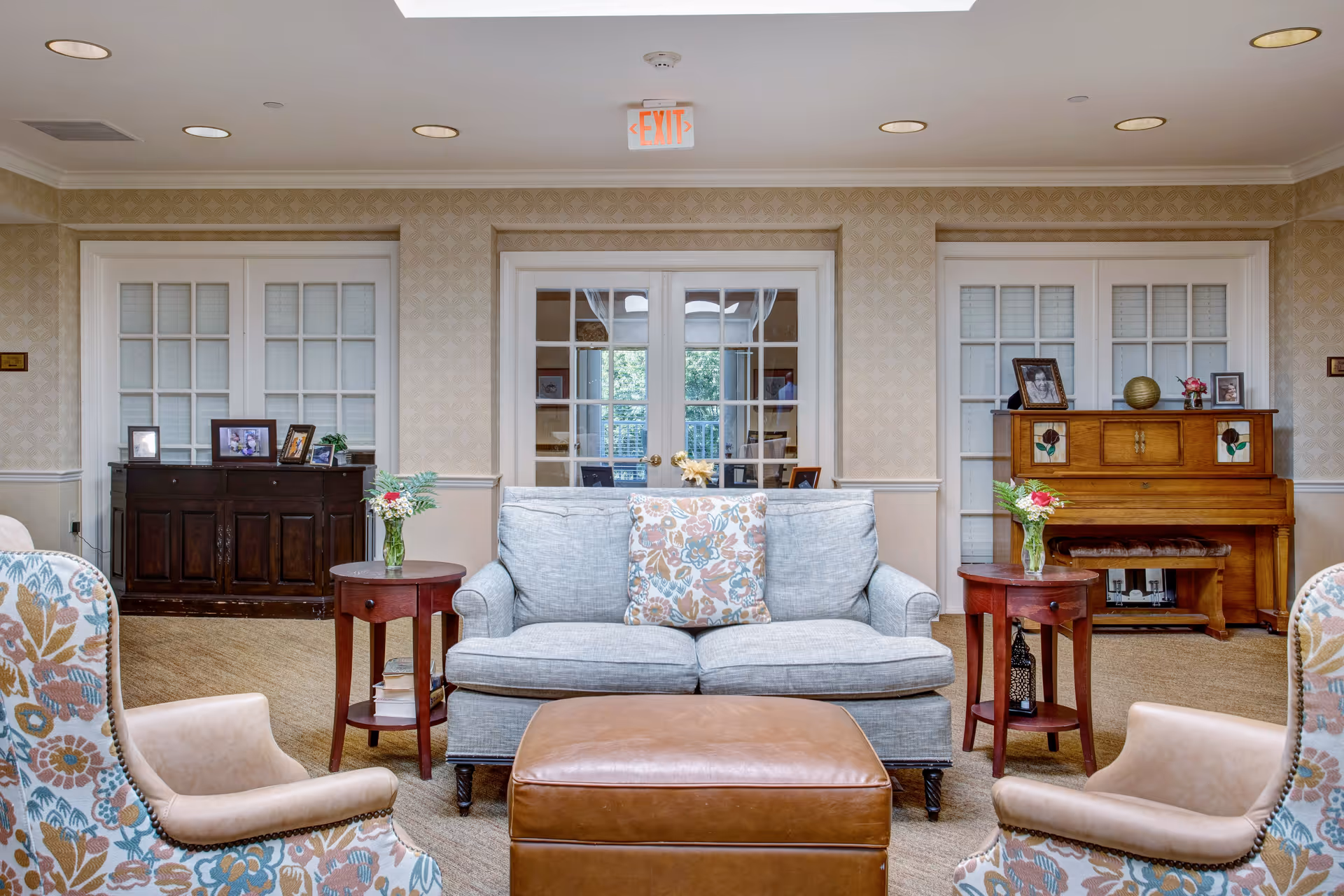 A cozy living room area with a light blue sofa adorned with a floral cushion, two floral patterned armchairs, and a brown leather ottoman in the center. There are two small wooden side tables with vases of flowers on either side of the sofa. In the background, there is a wooden piano with framed photos and decorative items on top, and a dark wooden cabinet with framed photos. The walls have a light patterned wallpaper, and there are glass-paneled doors behind the sofa.