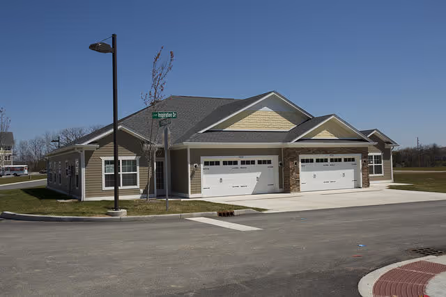 Exterior view of a single-story residential building with beige siding, stone accents, and two white garage doors under a clear blue sky. There is a street sign labeled 'Basingstone Dr' and a street lamp near the building.