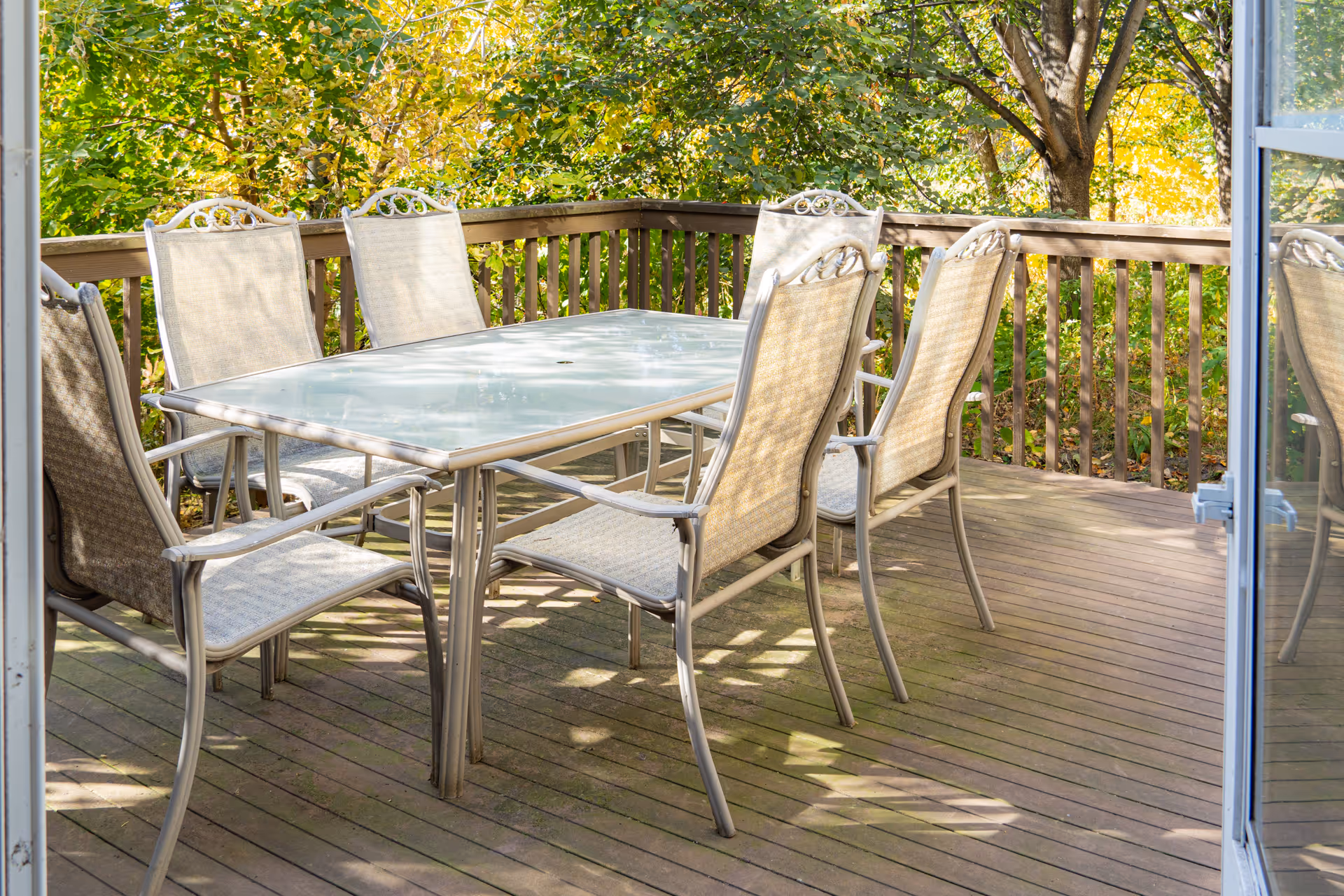 Outdoor wooden deck with a glass-top table and six beige mesh chairs surrounded by green leafy trees.