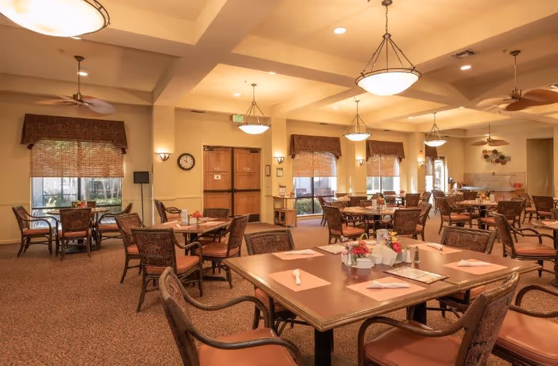 A spacious senior living dining room with multiple tables and chairs arranged neatly. Each table is set with placemats, napkins, and small flower arrangements. The room is warmly lit with ceiling lights and has large windows with woven shades allowing natural light to enter. The walls are painted in a soft beige tone, and there are ceiling fans and wall sconces for additional lighting.