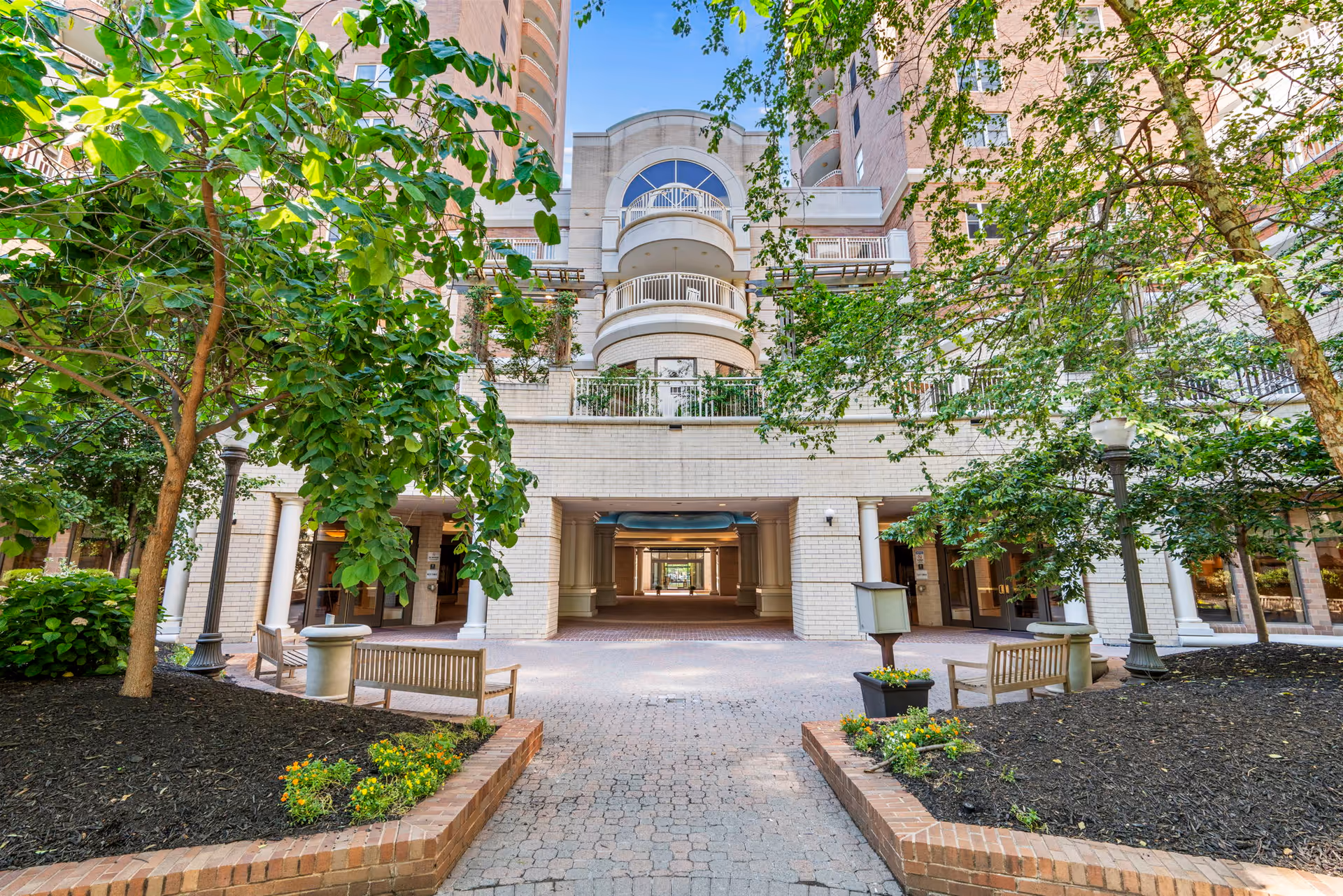 Outdoor courtyard area of a senior living facility named The Jefferson, featuring brick pathways, landscaped garden beds with trees and flowers, benches for seating, and a multi-story building with balconies in the background under a clear blue sky.