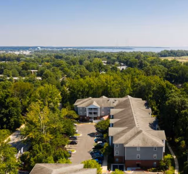 Aerial view of a multi-story senior living complex surrounded by trees with water visible on the horizon.