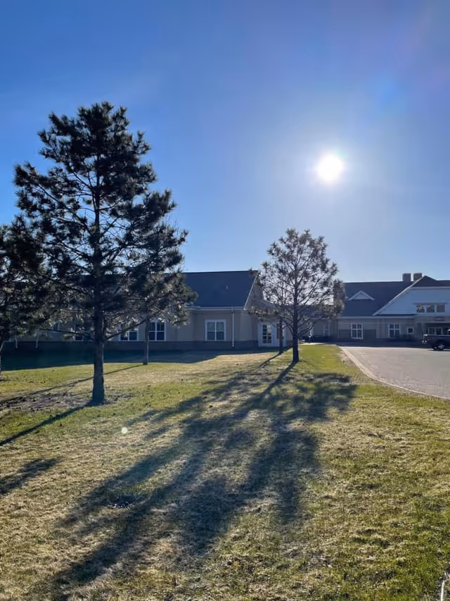 Sunlit front lawn with trees leading to the facade of a single-story senior living building under a clear blue sky.