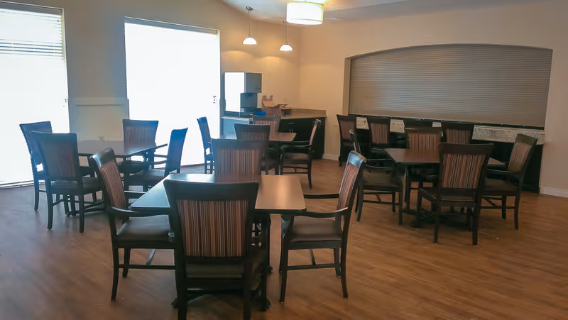 A dining room with multiple wooden tables and chairs arranged neatly on a wooden floor. The room has large windows with blinds, pendant lights, and a counter area with a coffee machine and other items.