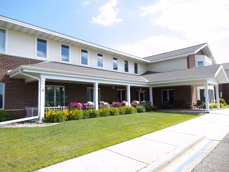 Exterior view of a two-story senior living facility building with brick and white siding. The building has a covered entrance supported by white columns, flower beds with colorful flowers, and a well-maintained lawn in front. The sky is partly cloudy.