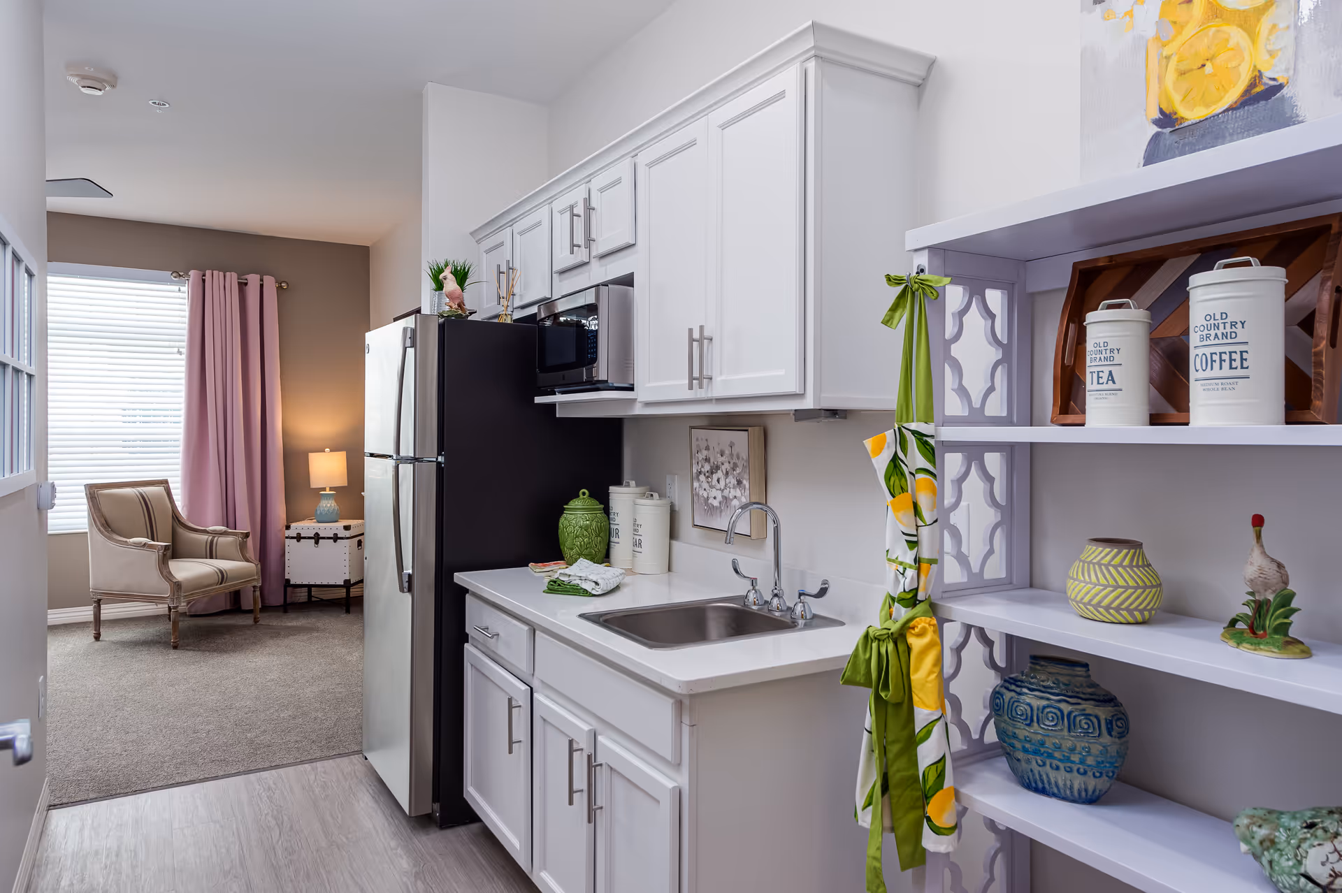 A small kitchen area with white cabinets, a stainless steel refrigerator, a microwave, and a sink. To the right, there is a white shelving unit with decorative items including canisters labeled tea and coffee, vases, and a small ceramic rooster. In the background, there is a cozy sitting area with a beige armchair, a side table with a lamp, and a window with pink curtains.