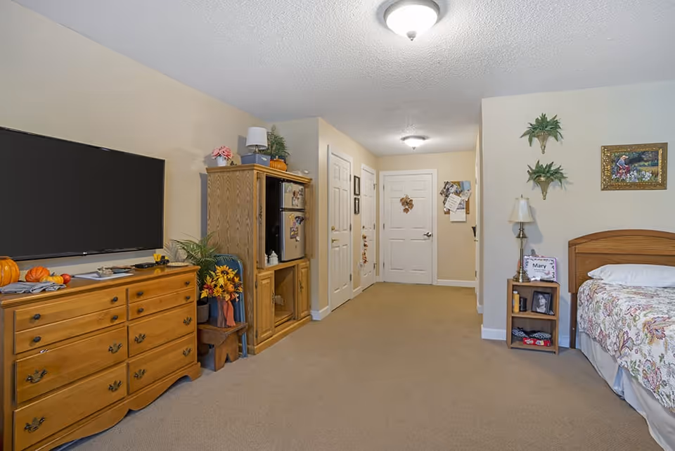 A cozy bedroom with beige walls and carpet. On the left side, there is a wooden dresser with a flat-screen TV mounted above it and some decorative pumpkins and flowers on top. Next to the dresser is a wooden cabinet with a small refrigerator and more decorations. On the right side, there is a wooden bed with a floral bedspread, a small nightstand with a lamp, framed photos, and wall-mounted plants and a painting above the bed. The room has two ceiling lights and several closed white doors along the back wall.