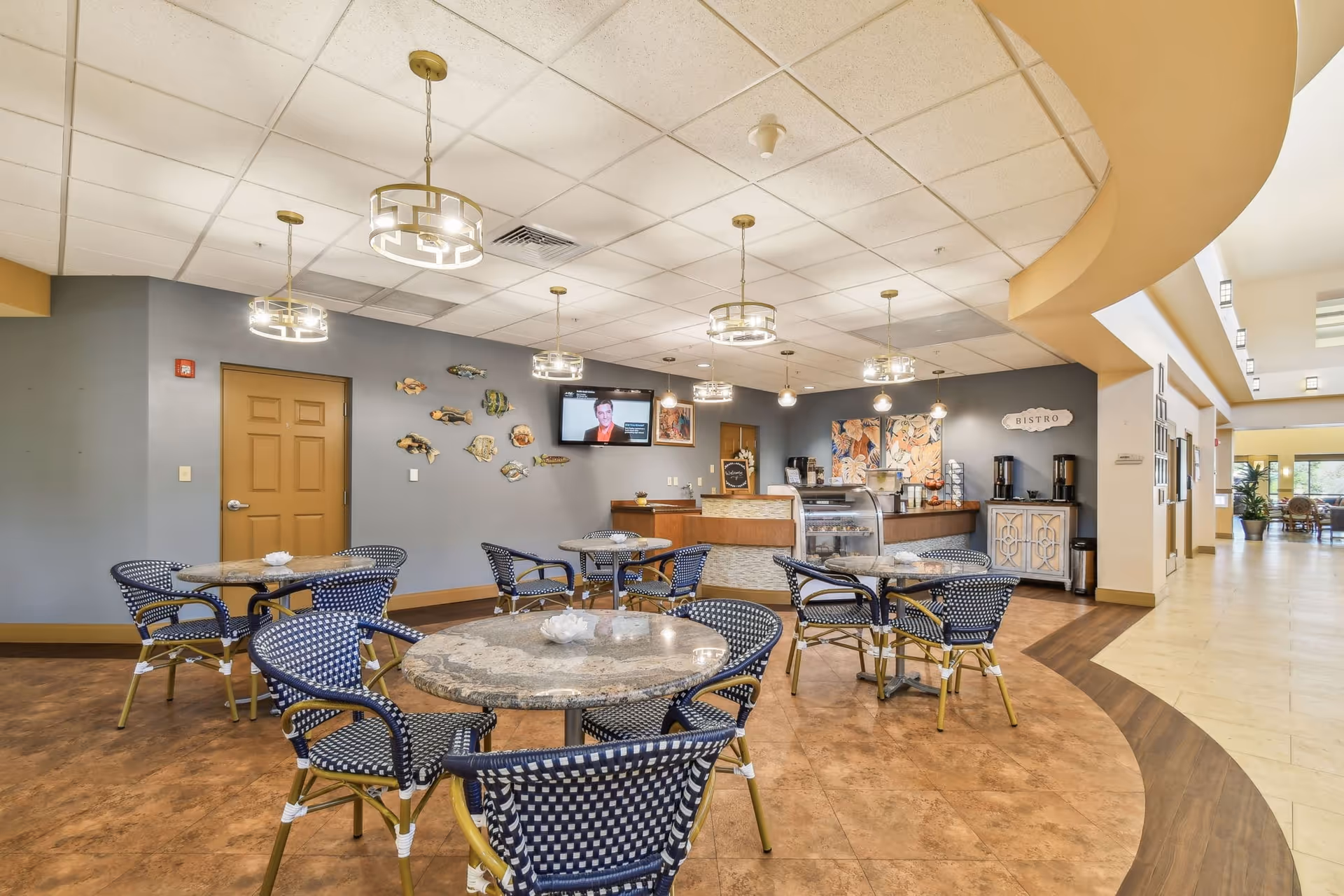 A bright and spacious dining area with several round tables and blue woven chairs. The room features a curved ceiling design with modern hanging light fixtures. On one side, there is a counter area labeled 'Bistro' with coffee machines and a display case. The walls are decorated with fish artwork and a mounted TV screen. The floor has a combination of tile and wood patterns, and a hallway extends to the right with additional seating visible in the distance.