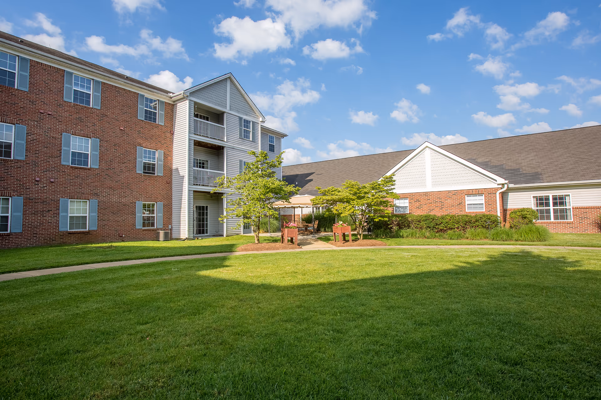 Exterior view of a senior living facility building with red brick and white siding, multiple windows, and a well-maintained green lawn with a paved walkway and small trees under a partly cloudy blue sky.