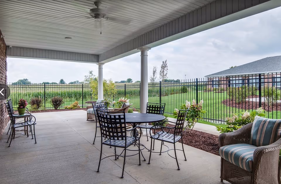 Covered outdoor patio area with metal table and chairs, cushioned wicker chair, and a bench. The patio overlooks a fenced grassy area with shrubs and trees under a cloudy sky.