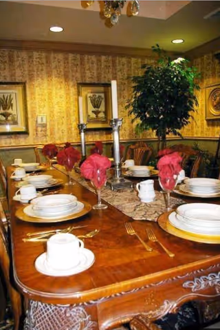 A formal dining room with a wooden table set for six people. The table is adorned with white plates, cups, gold chargers, and red cloth napkins folded in wine glasses. There are two tall silver candlesticks in the center and a decorative runner. The room has patterned wallpaper, framed botanical prints on the walls, and a large green potted plant in the corner.