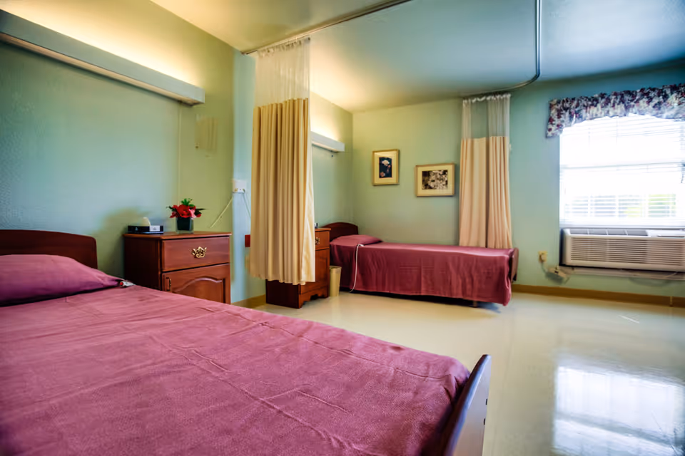 A shared bedroom in a skilled nursing facility with two single beds covered in maroon bedspreads. Each bed has a wooden nightstand beside it, one with a small vase of flowers and a box of tissues. There are privacy curtains between the beds, light green walls, two framed pictures on the wall, a window with floral valance, and an air conditioning unit below the window.