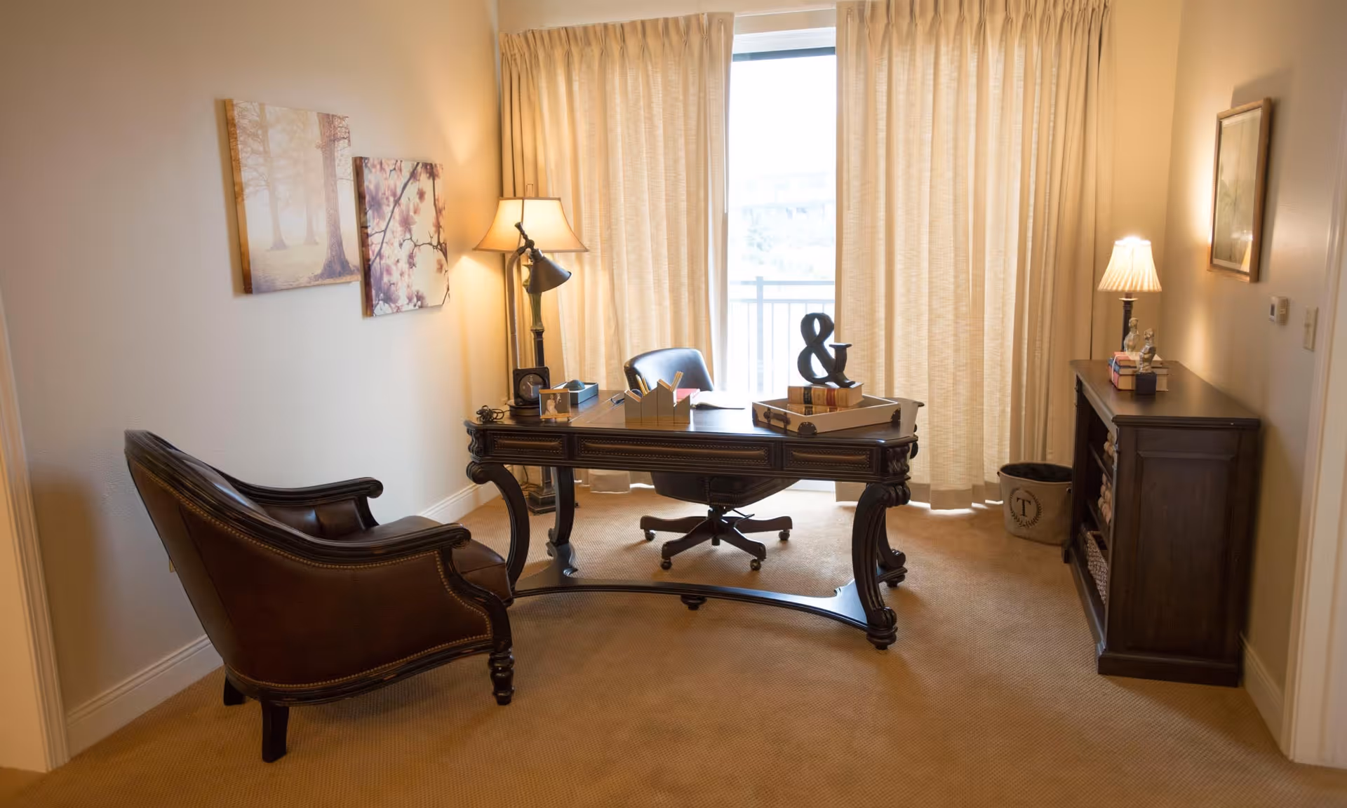 A cozy office room with a dark wooden desk and a leather chair. The desk has decorative items including books and an ampersand symbol. There is a floor lamp beside the desk and a wooden cabinet with a table lamp and books on the right side. Beige curtains cover a large window or sliding door in the background. Two framed pictures hang on the left wall.