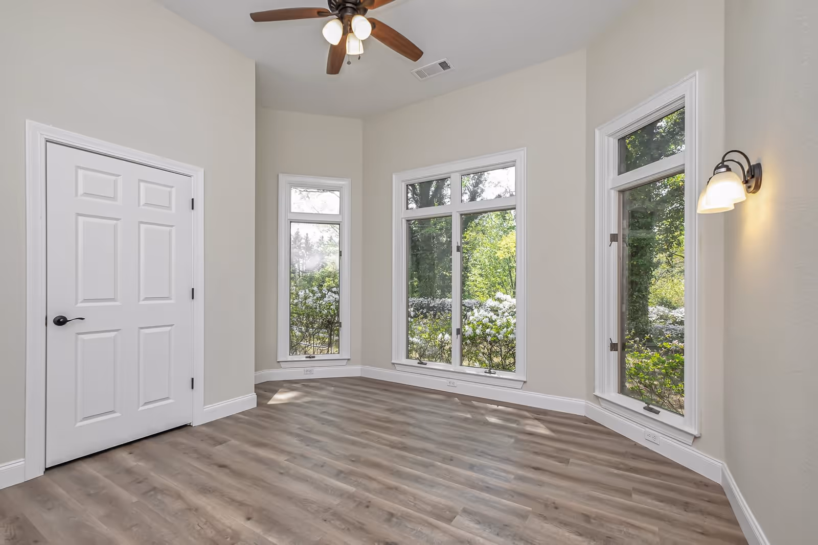 Empty bright room with wood-look flooring, three tall windows, a ceiling fan, a wall sconce, and a white door.