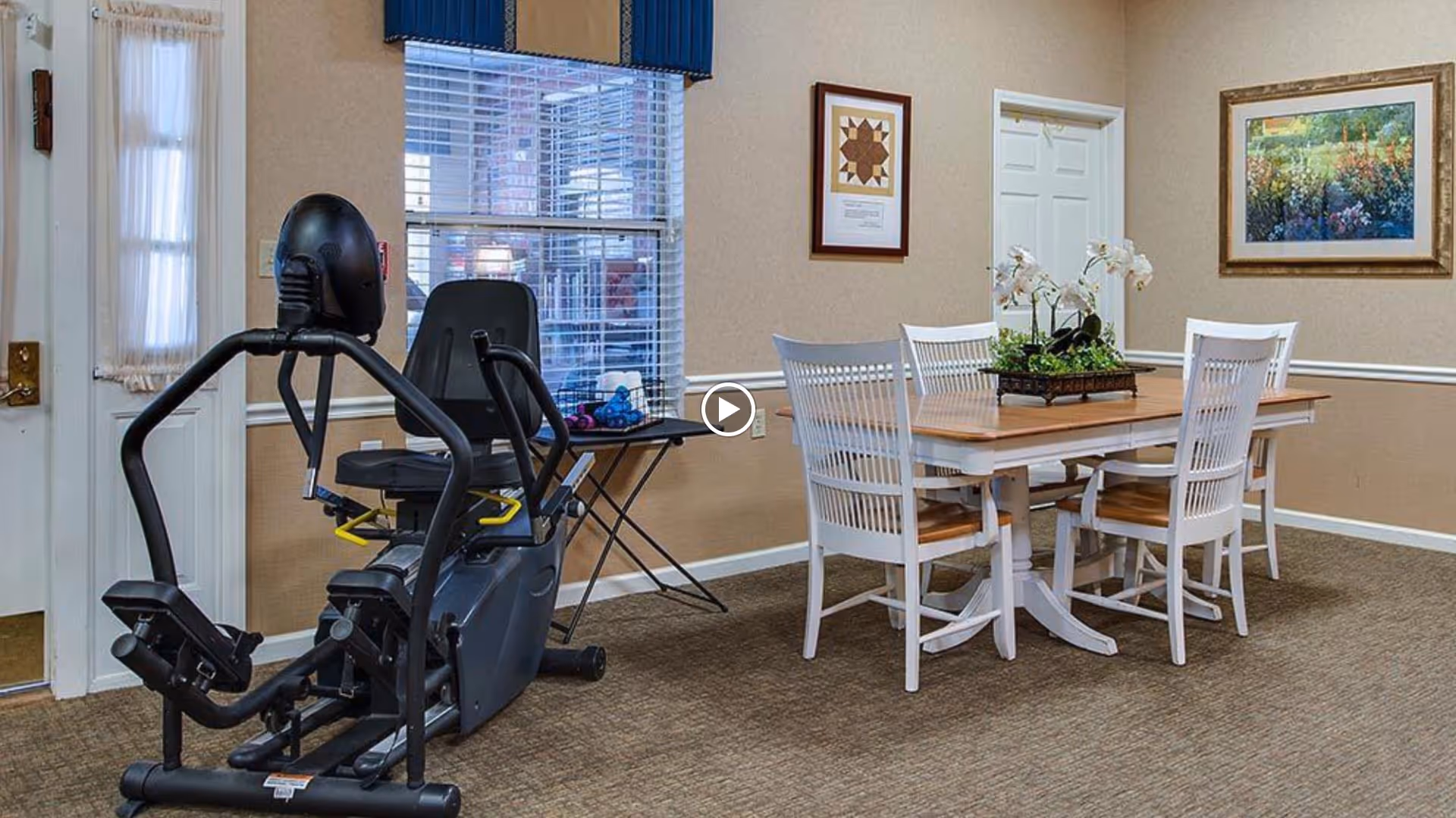 Interior room with a recumbent exercise bike near a window with blinds and a small folding table. A wooden dining table with four white chairs is positioned against the wall, decorated with a floral centerpiece. The walls are beige with white trim, and two framed pictures hang on the walls.
