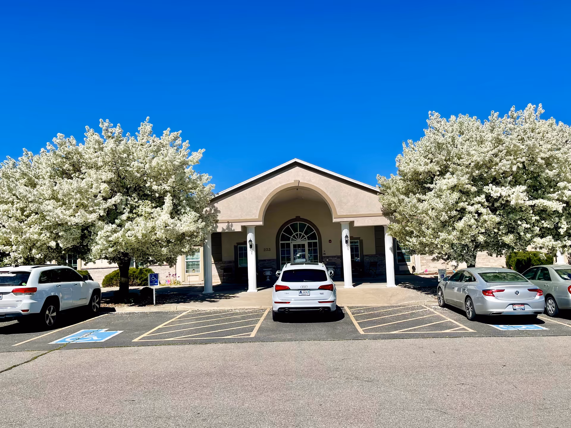 Front entrance of a single-story assisted living facility with a covered portico, parked cars and flowering trees under a clear blue sky.