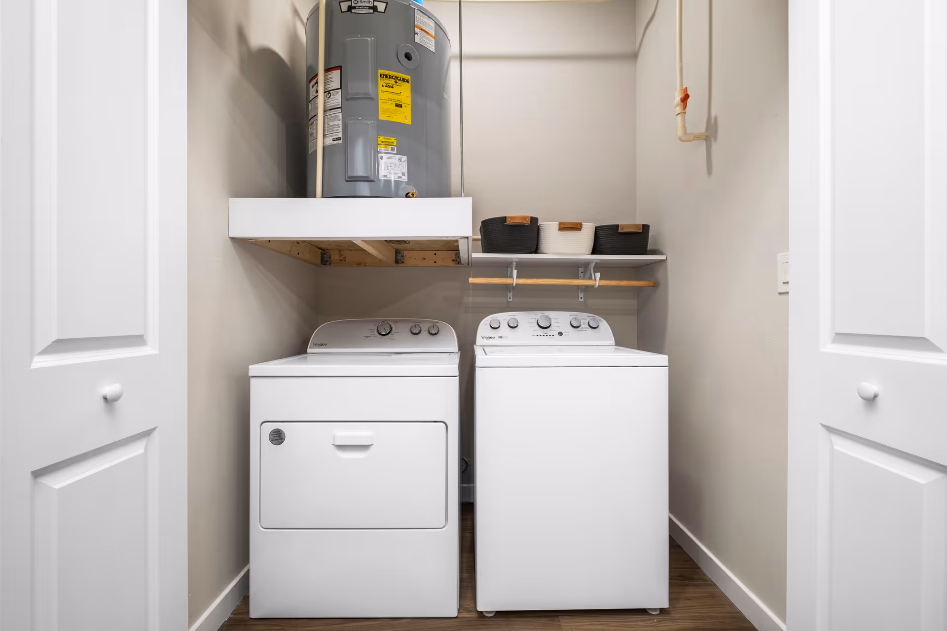 A laundry room with a white washing machine and dryer side by side. Above the machines, there is a shelf holding three storage baskets and a water heater mounted on a platform. The room has beige walls and wooden flooring, with white double doors partially open.