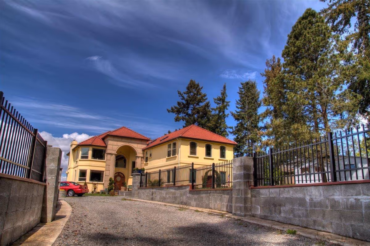 View of a large, two-story yellow building with a red tiled roof, surrounded by a black metal fence and concrete wall. There is a gravel driveway leading up to the entrance, with a red car parked on the left side. Tall trees are visible behind the building under a partly cloudy blue sky.