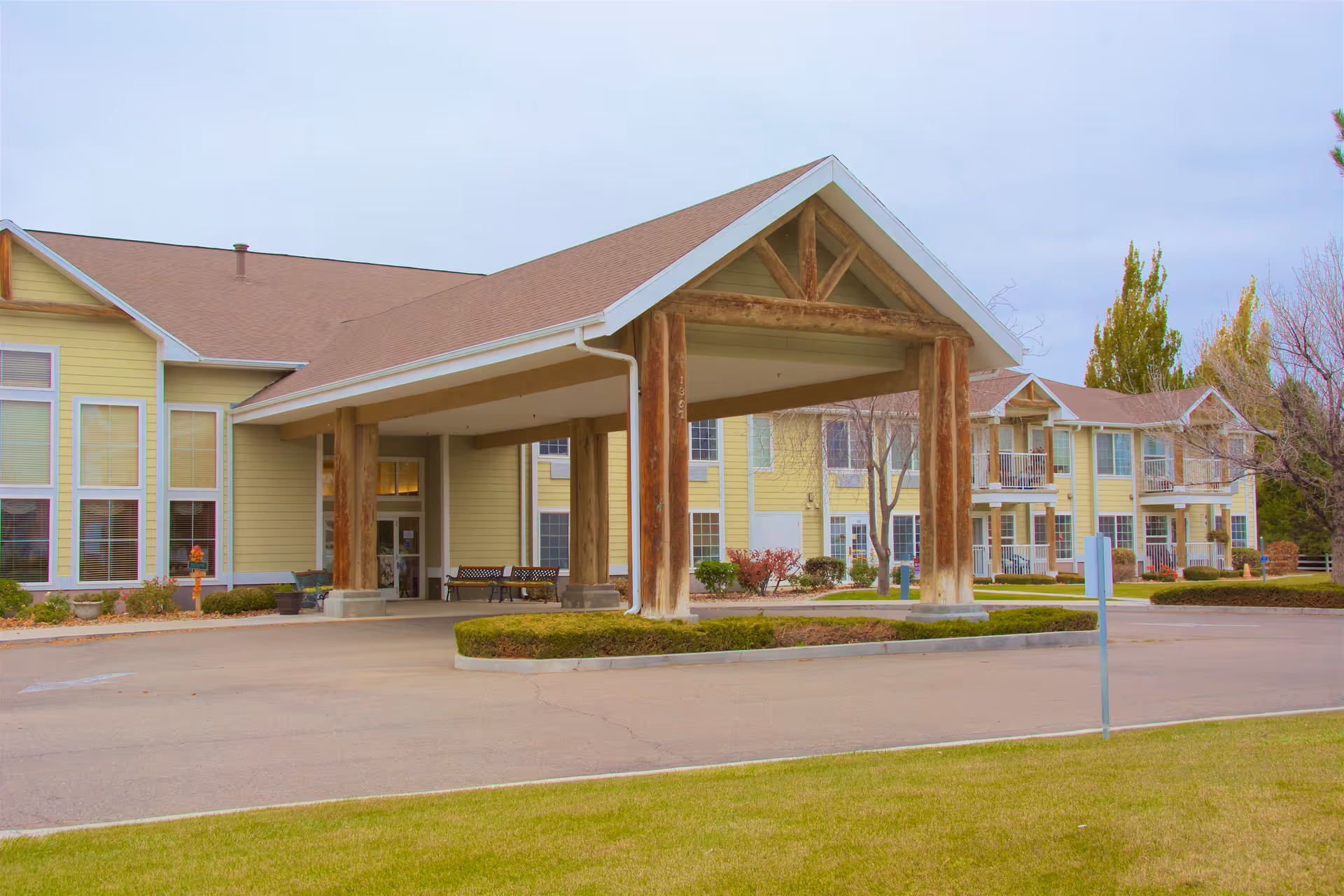 Front entrance of a yellow senior living facility with a covered porte-cochere, balconies, and a landscaped driveway.