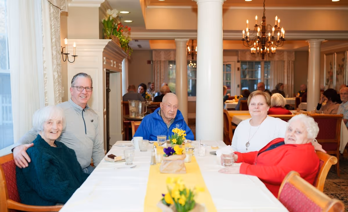 A group of five elderly people sitting around a dining table in a well-lit dining room with white tablecloths and yellow table runners, decorated with small flower arrangements. The room has large windows with curtains, white columns, and chandeliers. Other people are visible dining in the background.