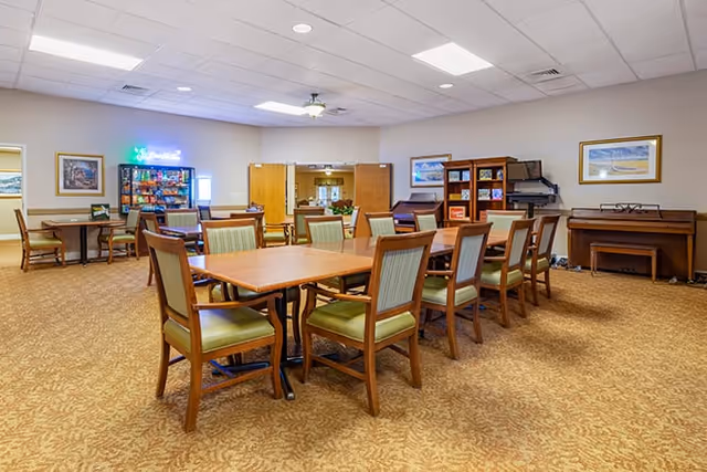 A spacious common area in a senior living facility with multiple wooden tables and green cushioned chairs arranged for group seating. The room has beige carpet, light-colored walls adorned with framed artwork, a piano against one wall, bookshelves, and a vending machine in the corner. The ceiling has recessed lighting and a ceiling fan.