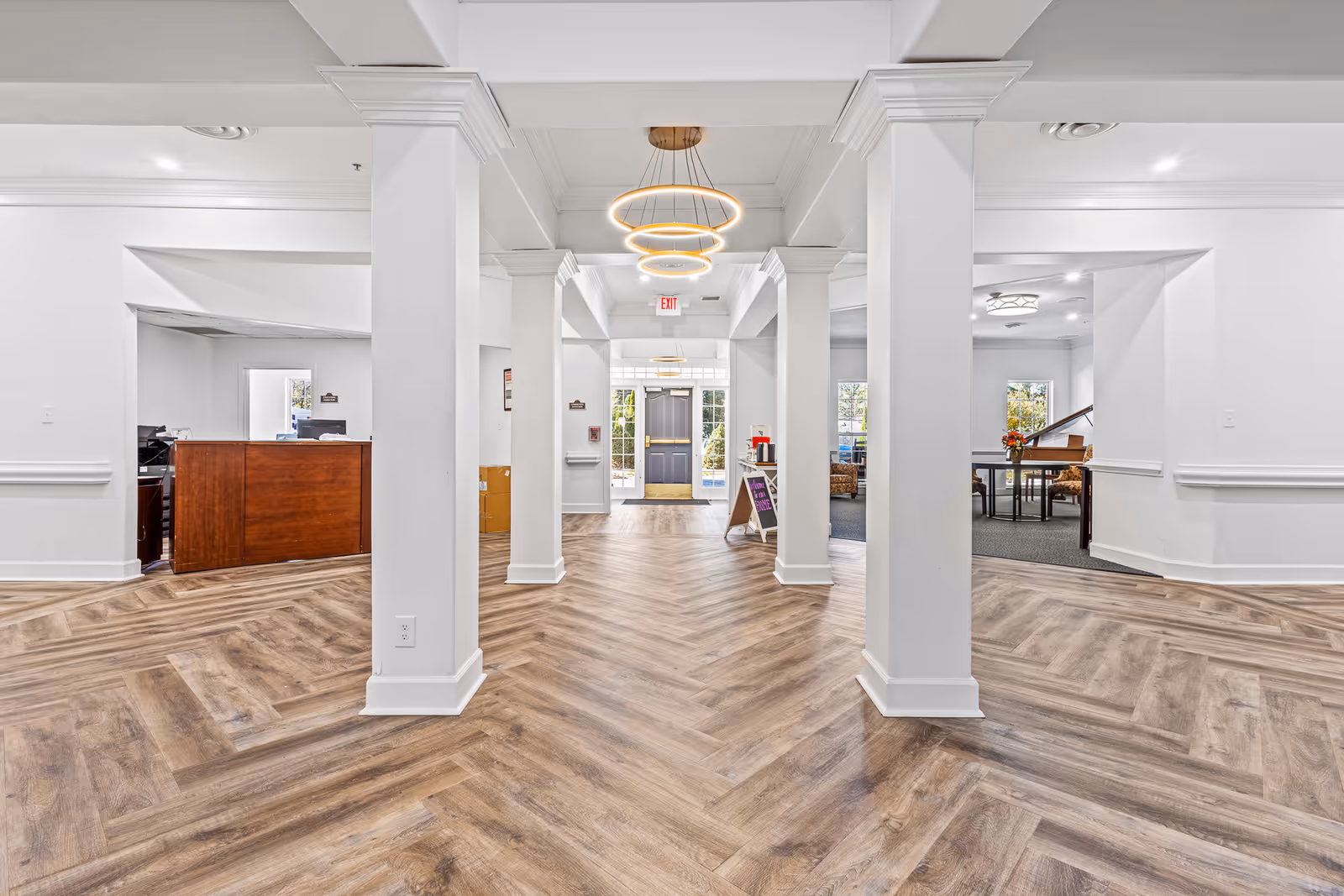 Bright and spacious interior hallway of a senior living facility with white walls, wooden herringbone-patterned flooring, and modern circular ceiling lights. The hallway features multiple white columns and leads to a glass door exit. On the left side, there is a wooden reception desk, and on the right side, a sitting area with a piano and chairs is visible.