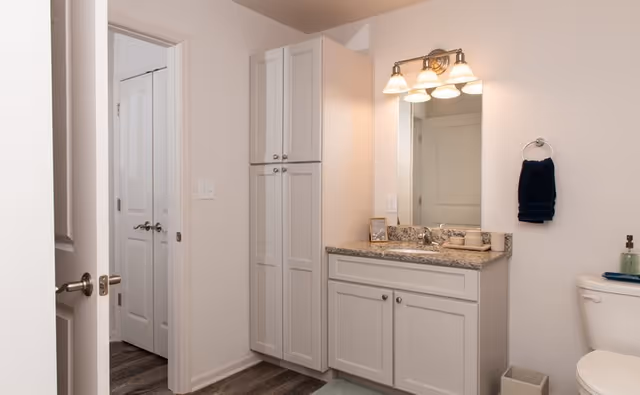 A clean and modern bathroom featuring white cabinetry with a granite countertop, a large mirror with three-light fixture above, a towel ring with a dark blue towel, a white toilet, and a small trash bin. The floor has wood-style flooring and there are white doors visible in the background.