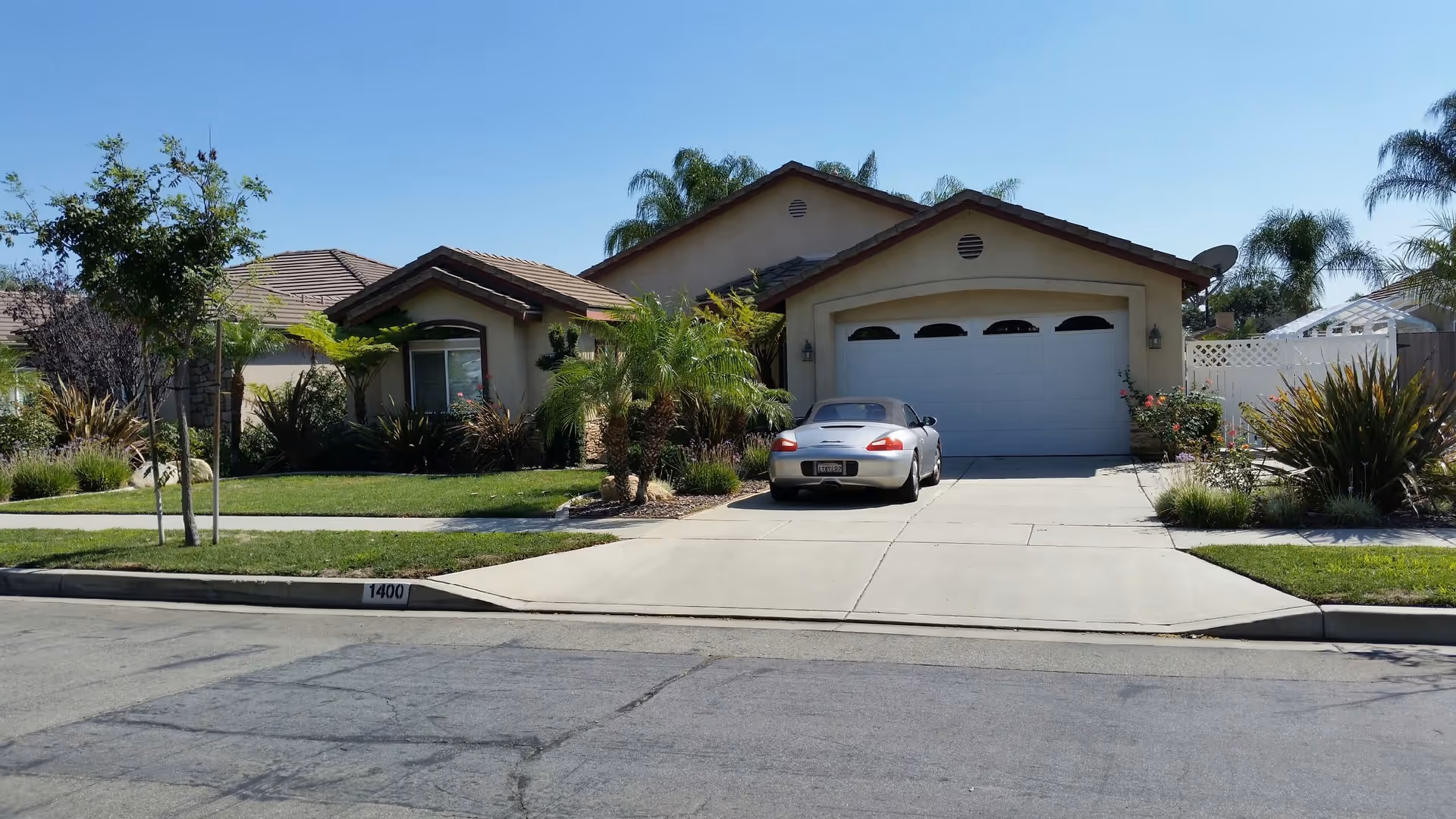 Front exterior view of a single-story house with a tiled roof, a two-car garage, a driveway with a silver convertible car parked, and a well-maintained front yard with green grass, palm trees, and other plants under a clear blue sky.
