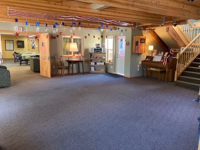 Large open communal lounge with carpeted floor, piano, seating, staircase, and patriotic star decorations hanging from the ceiling.