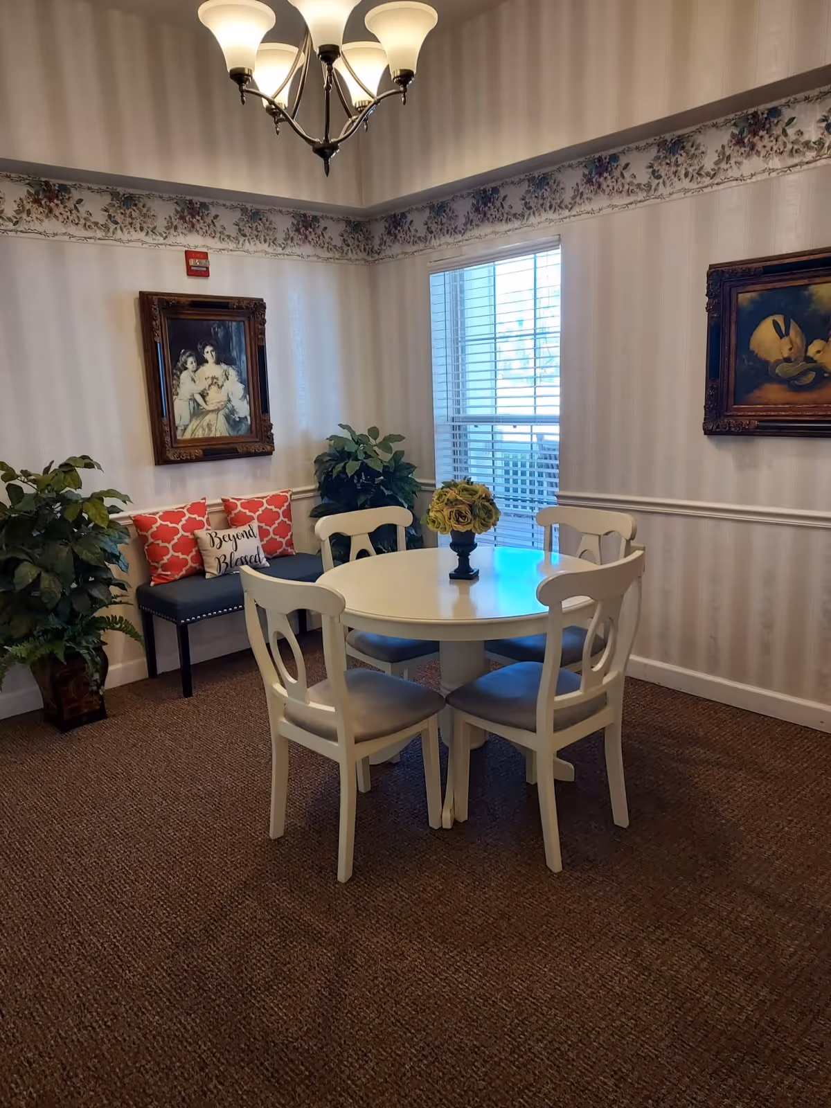 A cozy dining nook with a round white table and four chairs, potted plants, framed paintings, and a bench with decorative pillows under a chandelier.