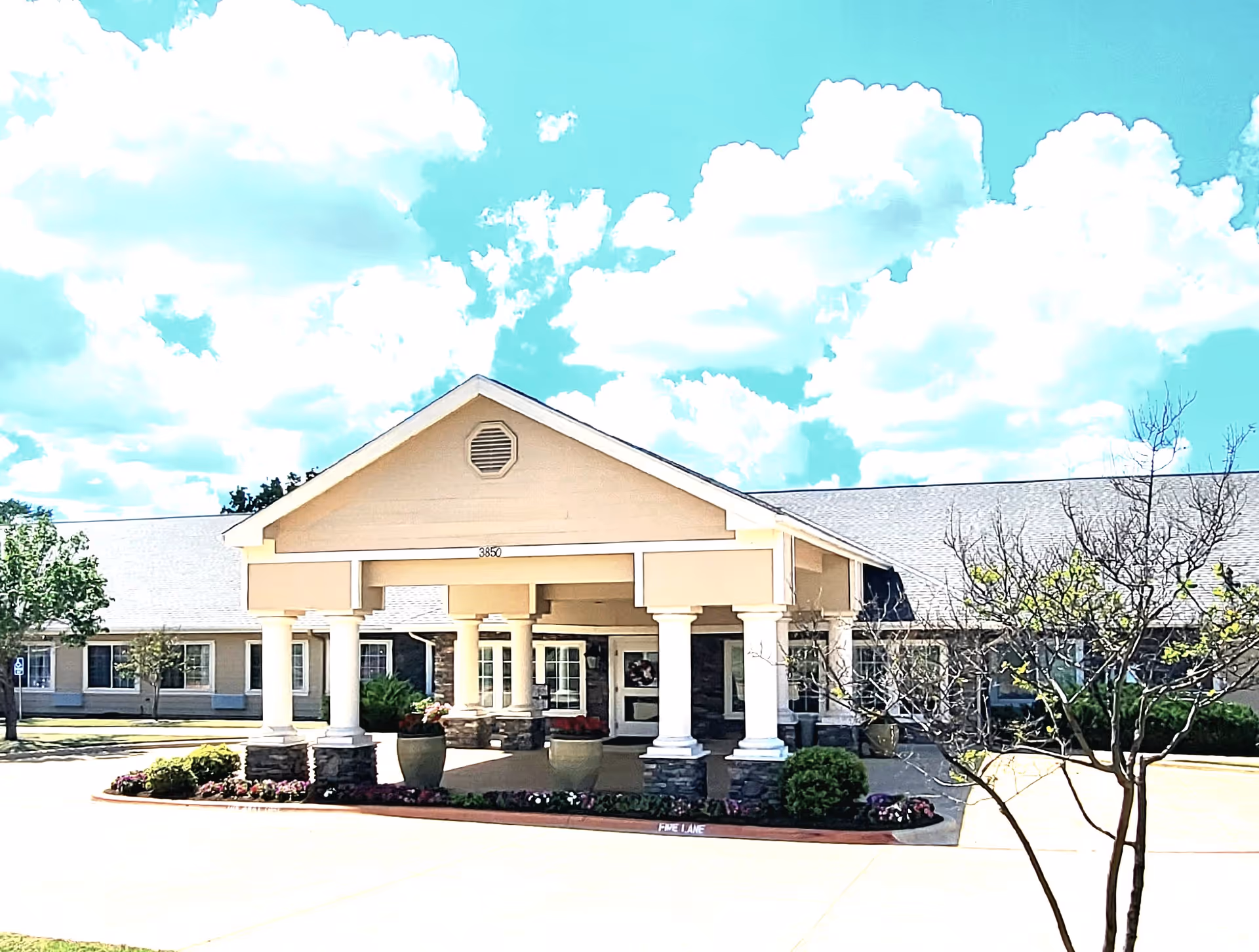 Front exterior view of Hudson Creek Alzheimer's Special Care Center showing a covered entrance with white columns and stone bases, landscaped flower beds, and a clear blue sky with clouds.