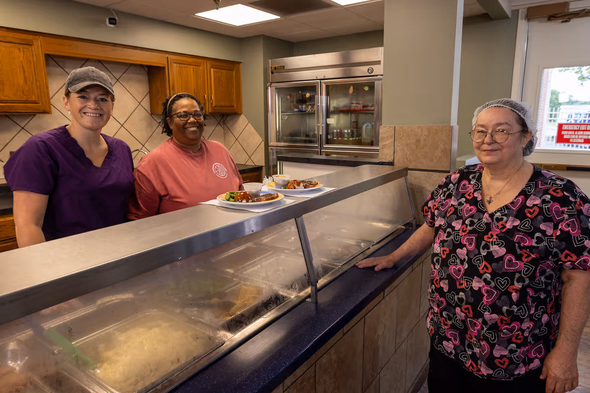 Three women standing behind a food serving counter in a kitchen area. Two women are behind the counter smiling, one wearing a purple scrub top and a cap, the other wearing a red shirt and glasses. The third woman, wearing a patterned scrub top and a hairnet, stands in front of the counter. Plates of food are placed on the counter. The background shows wooden cabinets, a tiled wall, and a refrigerator.