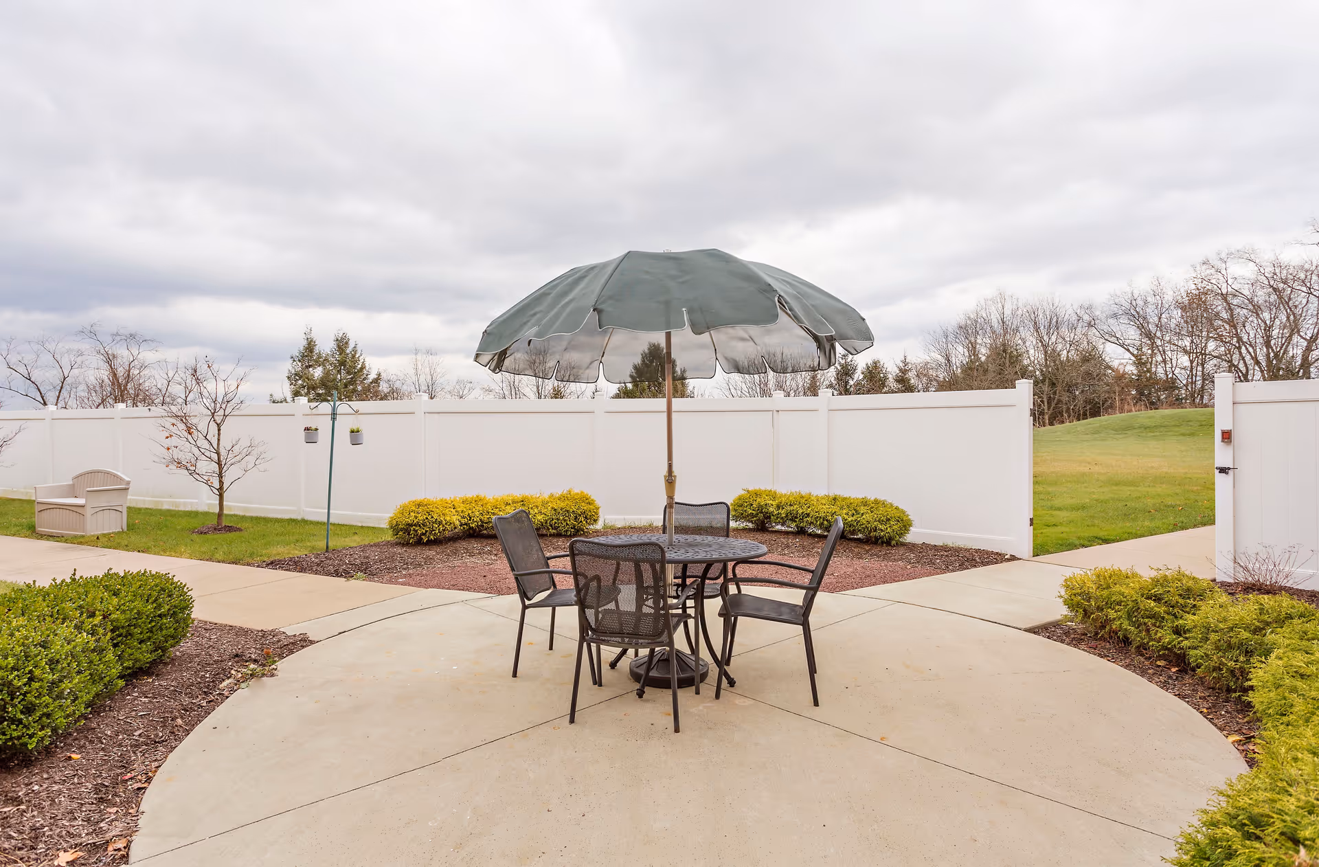 Outdoor patio area with a round metal table and four matching chairs under a large green umbrella. The patio is surrounded by neatly trimmed bushes and a white privacy fence, with a cloudy sky overhead.