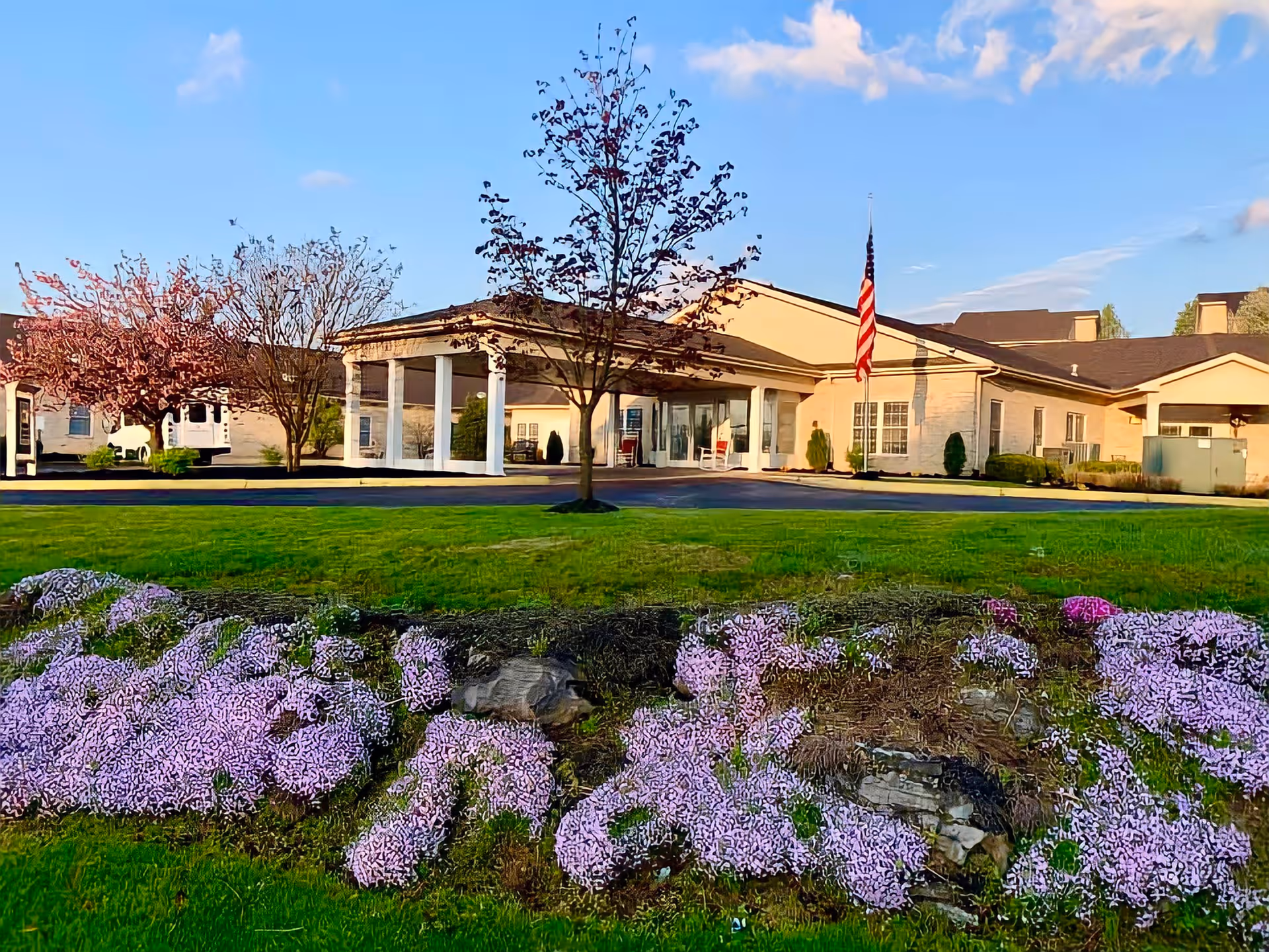 Exterior view of The Maynard facility showing a single-story building with a covered entrance supported by white columns. There is a well-maintained lawn with purple flowers in the foreground, a tree in the center, and an American flag near the entrance. The sky is clear with a few clouds.
