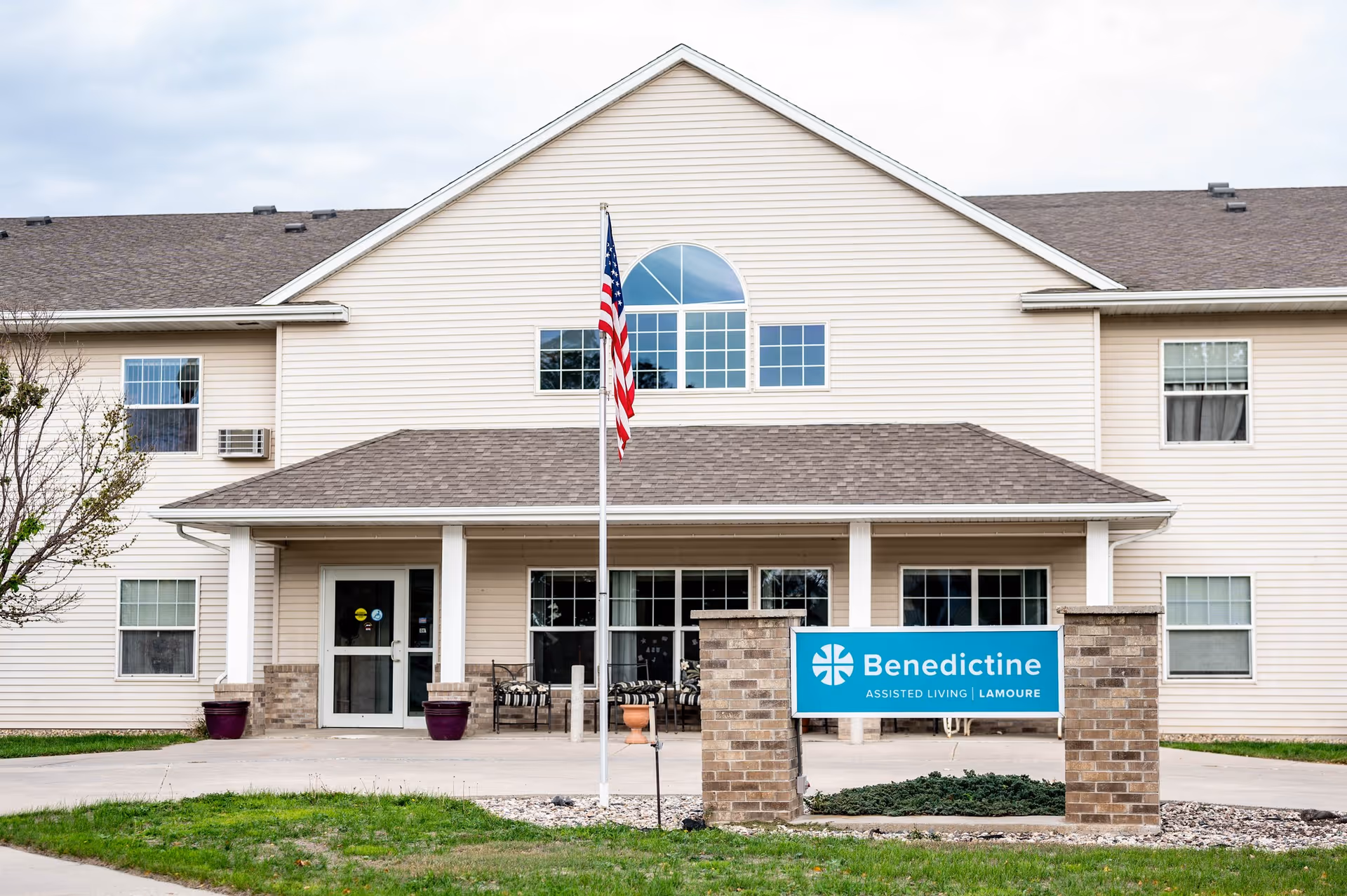 Front exterior view of the Benedictine Living Community-LaMoure building with a beige facade, multiple windows, a covered entrance with seating, an American flag on a pole, and a blue sign displaying the facility name.