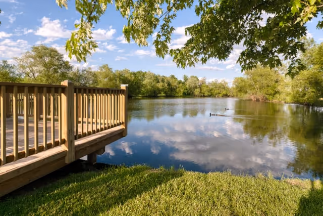 A peaceful outdoor scene featuring a wooden deck overlooking a calm pond surrounded by green trees under a partly cloudy blue sky. Two ducks are swimming on the pond, and green grass is visible in the foreground.