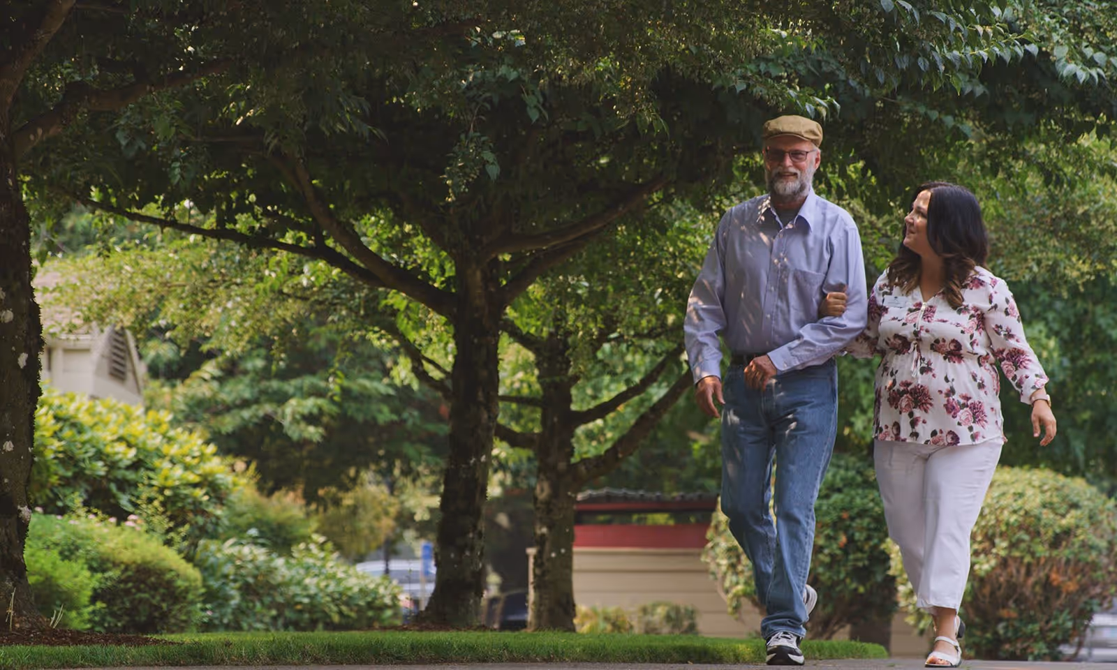 An elderly man wearing a cap, glasses, and a light purple shirt walks arm-in-arm with a woman in a white floral blouse and white pants along a shaded path surrounded by green trees and bushes.